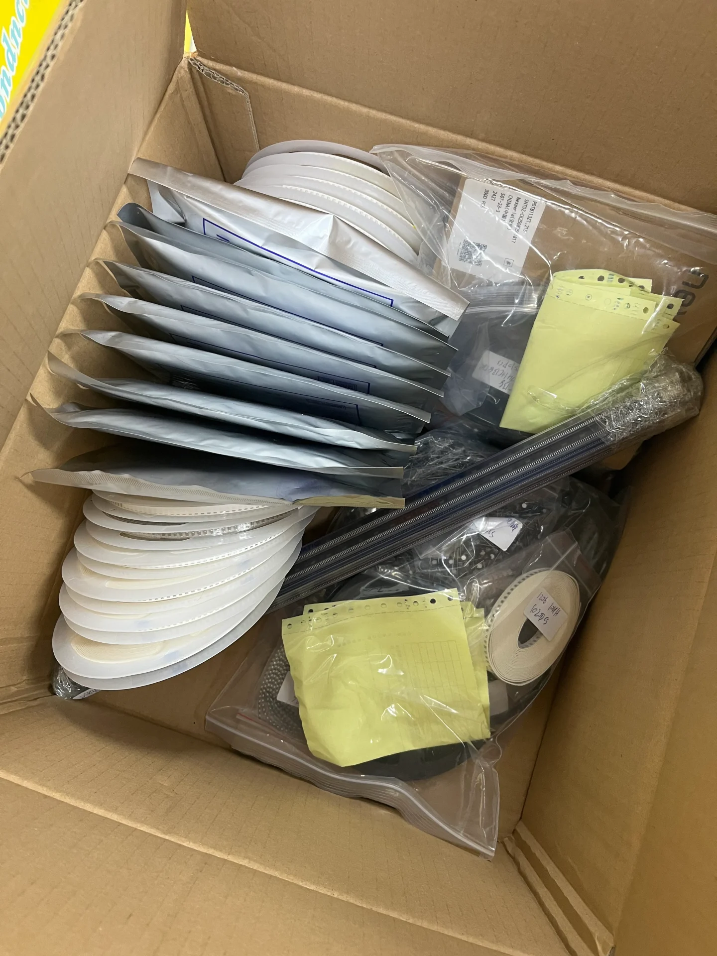Open cardboard box containing mixed electronic components: tape reels, tube packaging, and individual bags for a complete BOM order